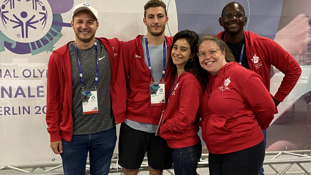Auf dem Foto sind fünf corporate Volunteers zu sehen, die lächelnd vor einer gebrandeten Wand mit Logo der Nationalen Spiele stehen. Alle tragen die Volunteer-Uniform und sehen glücklich aus.