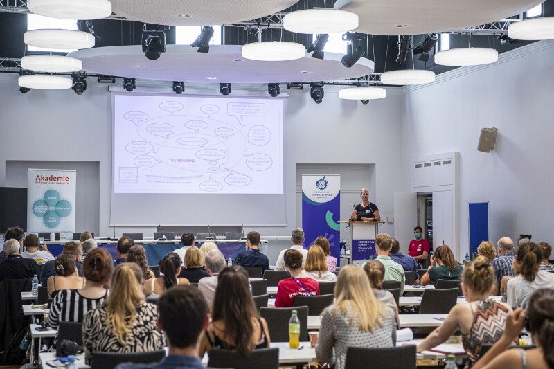 Das Foto zeigt viele Menschen, die einer Person zuhören. Eine Frau präsentiert und steht an einem Rednerpult.