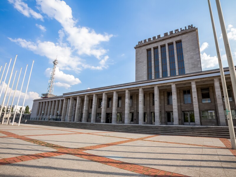 Das Foto zeigt einen gepflasterten Vorplatz und ein Gebäude mit Säulen auf dem Messe Berlin steht.