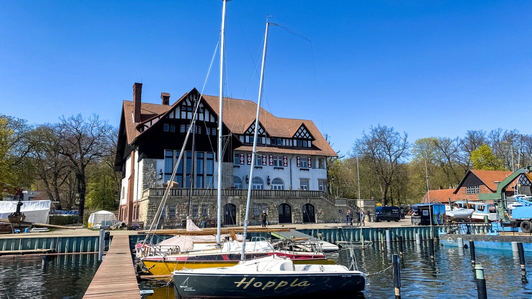 The photo shows a jetty with sailboats in the foreground and a white half-timbered house with natural stones at the base in the background.
