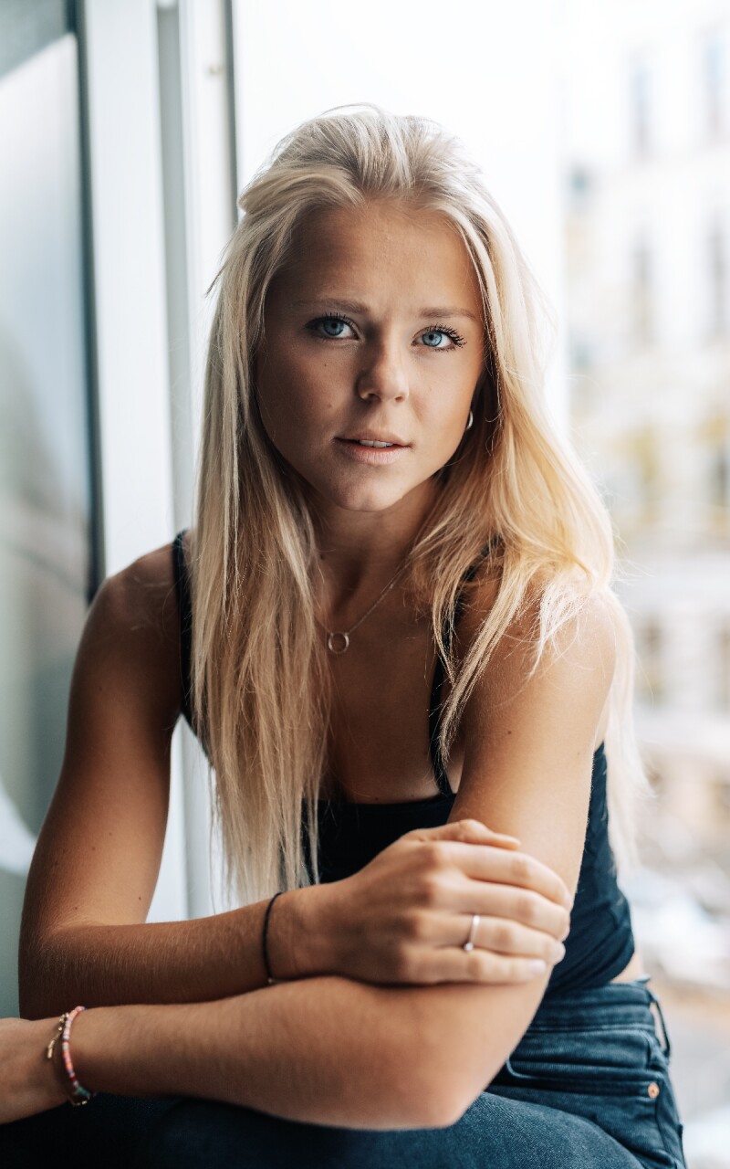 The photo shows the German running influencer Maren Schiller. She has long blonde hair, sits relaxed at a window and looks into the camera. 