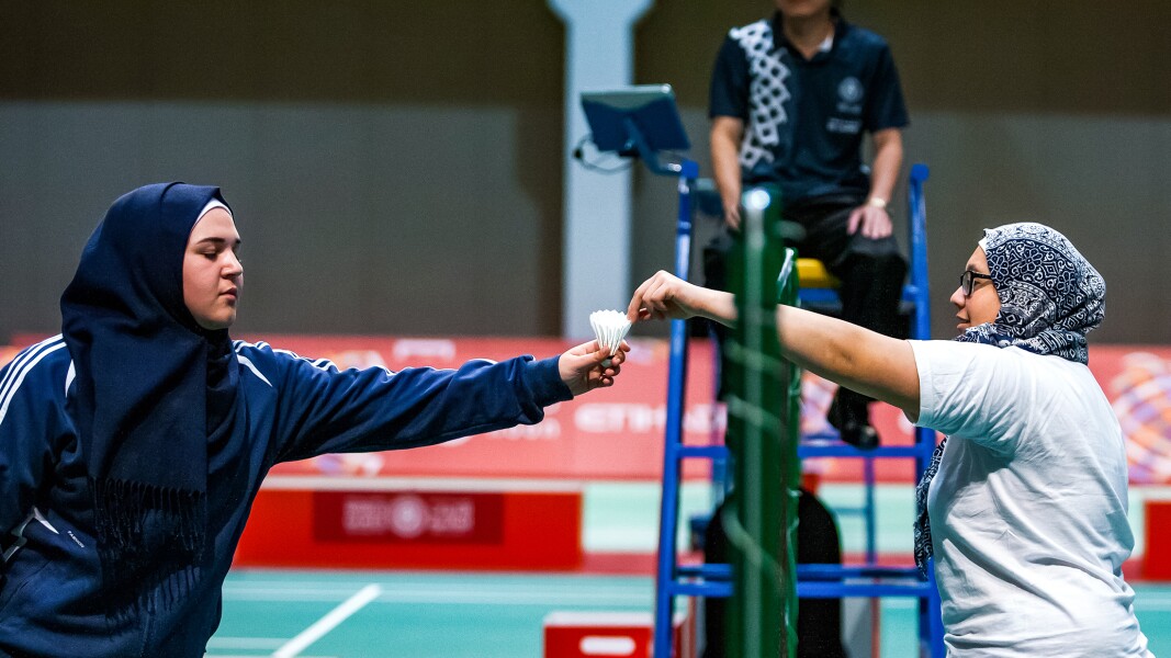Das Foto zeigt zwei Athletinnen beim Badminton, die sich gerade einen Federball über das Netz reichen.
