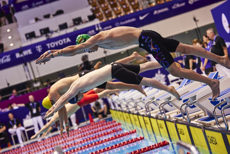 The photo shows the start of a swim race, with several athletes in different swimsuit colors taking a dynamic dive into the water.