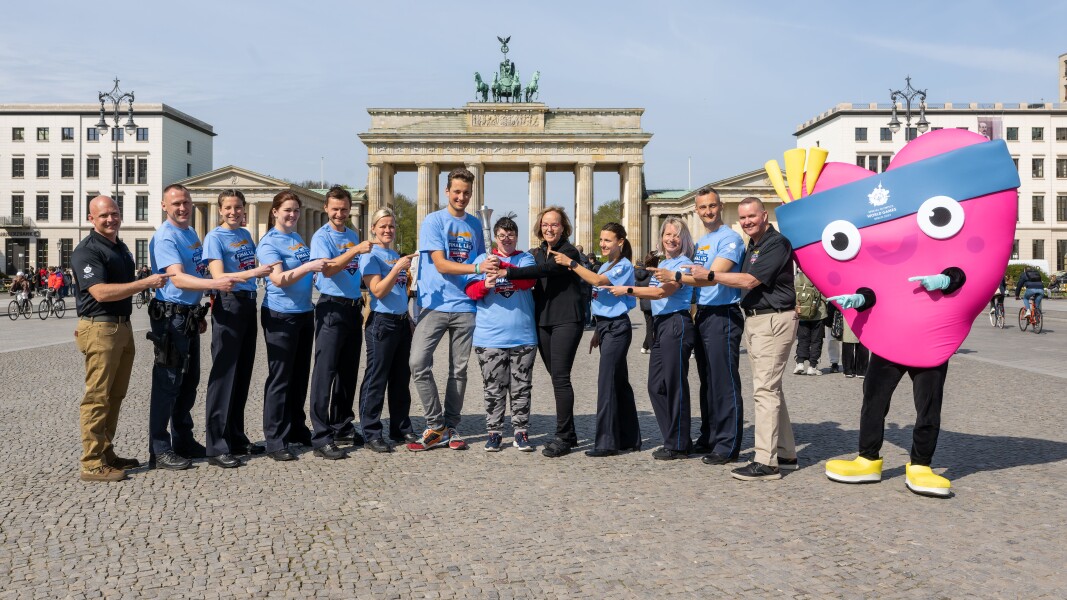 Auf dem Foto sind Polizisten und Athlet*innen zu sehen. Auch das Maskottchen Unity ist dabei. Sie sehen alle freudig aus und zeigen auf die Fackel.