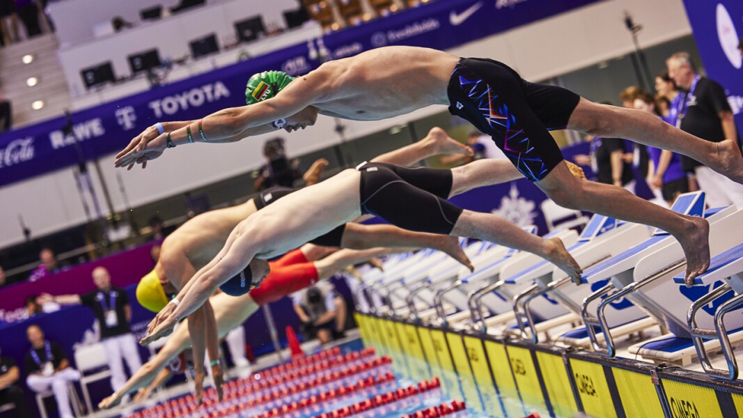 Das Foto zeigt den Start eines Schwimmrennens, bei der mehrere Sportler in unterschiedlichen Badehosen-Farben zum dynamischen Hechtsprung ins Wasser ansetzen.