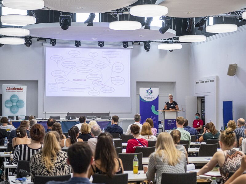The photo shows many people listening to one person. A woman is presenting and standing at a lectern.