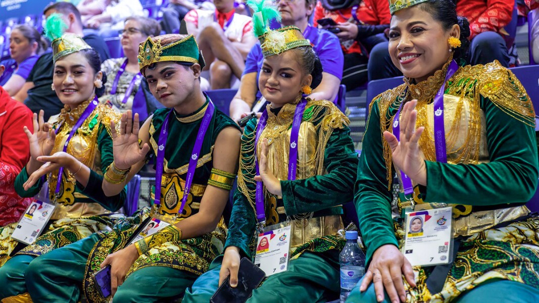 Auf dem Bild sieht man Athlet*innen und Zuschauende, die lächelnd in die Kamera winken.