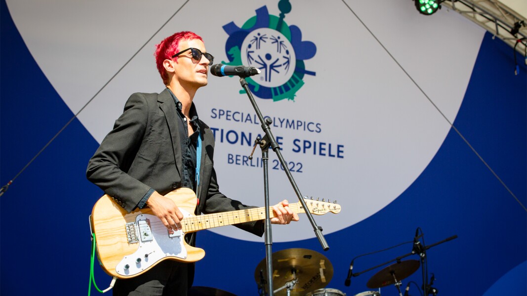 The photo shows a person on a stage holding a guitar and singing into a microphone. In the background you can see the logo of the Special Olympics National Games Berlin 2022.