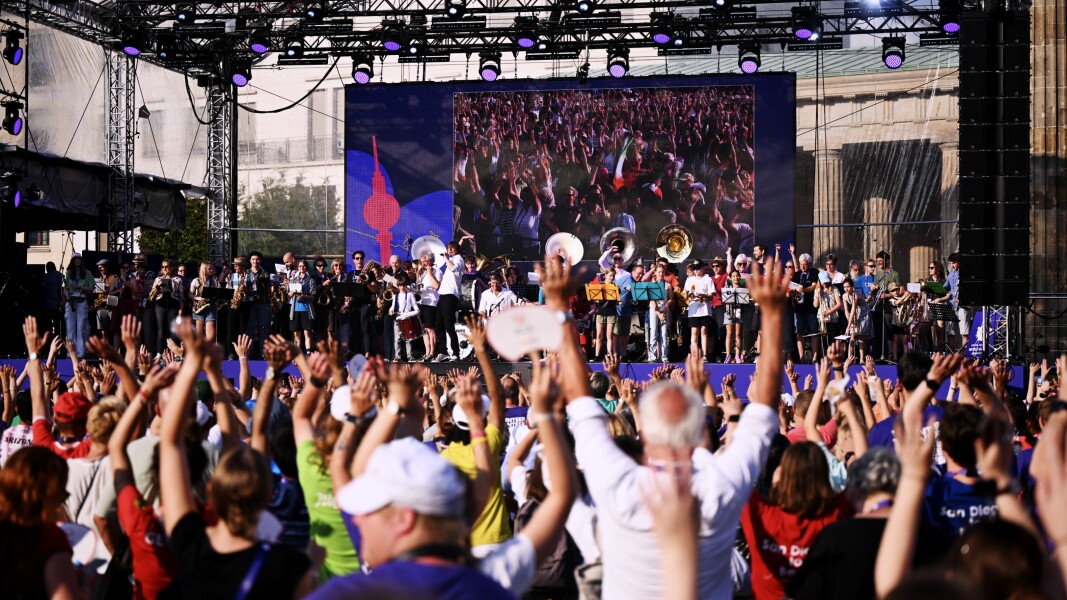 The photo shows the cheering crowd with arms raised at the closing ceremony of the World Games in Berlin. An orchestra is playing on the stage.