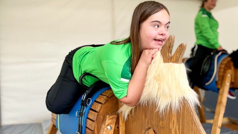 The photo shows a young girl at the competition free offer. She is sitting on a wooden horse and smiling towards the side.