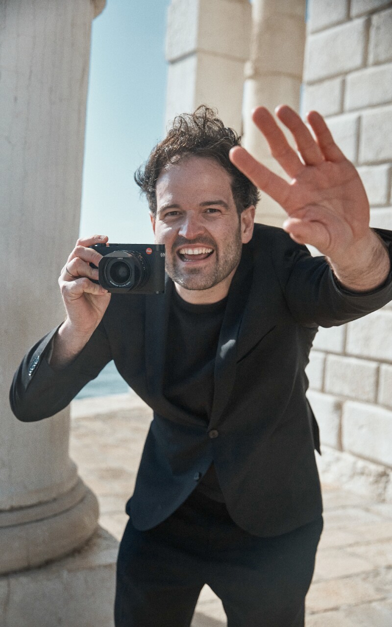 Das Foto zeigt den Fotografen Mike Meyer. Er winkt in Richtung Kamera und hält selbst auch eine Kamera in seinen Händen, dabei lacht er.