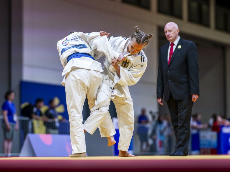 Zwei  Judo-Kämpferinnen ringen miteinander unter Beobachtung des Schiedsrichters.