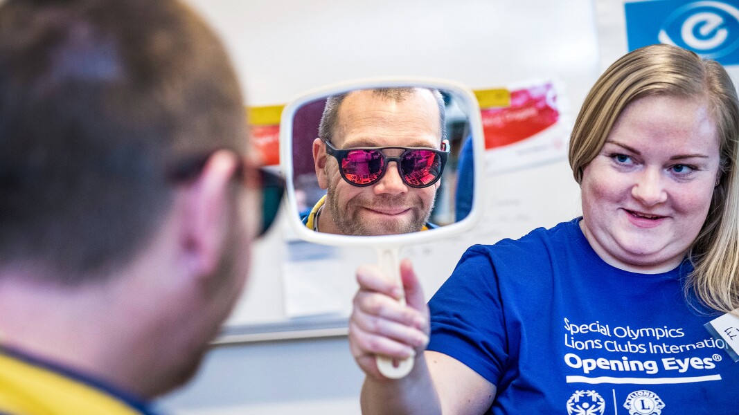 Das Foto zeigt eine Szene aus einer Healthy Athletes "Opening Eyes" Augenuntersuchung. Eine Frau hält einen Spiegel hoch und ein Mann sieht sich darin mit einer Sonnenbrille auf und lächelt.